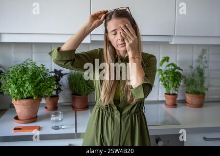 Schläfrige müde Frau, die an Schlaflosigkeit leidet, reibt am frühen Morgen die Augen auf die Küche mit Zimmerpflanzen. Stockfoto