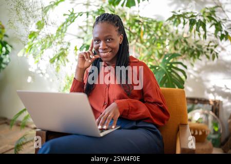 Glückliche, zufriedene schwarze Frau mit Laptop auf den Runden, die auf einem Stuhl in einem sonnigen Zimmer sitzt und lächelnd in die Kamera blickt. Stockfoto