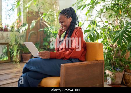 Fröhliche schwarze Frau mit Laptop lächelndem Blick auf den Bildschirm empfing gute Nachrichten im urbanen Dschungelraum sitzen. Stockfoto