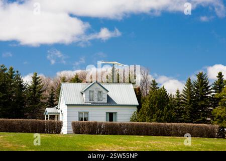 Weißes Haus mit blauem Dach auf einem grasbewachsenen Hügel. Das Haus ist von einer Hecke umgeben und auf dem Dach befindet sich eine kleine Antenne. Der Himmel ist klar und Stockfoto