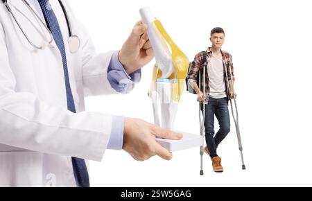 Männlicher Student, der mit Krücken geht, und Arzt, der ein Knieknochenmodell auf weißem Hintergrund hält Stockfoto