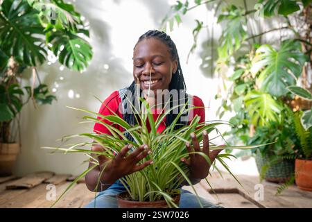 Fröhliche, glückliche afroamerikanische Frau hält Chlorophytum umgeben von Blumen und Pflanzen im Garten Stockfoto