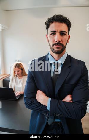 Ein entschlossener Geschäftsmann steht mit gekreuzten Armen und strahlt Vertrauen und Stolz aus, während sein Kollege intensiv an einem Laptop arbeitet und die Essenz von Teamarbeit und Professionalität am Arbeitsplatz verkörpert. Hochwertige Fotos Stockfoto
