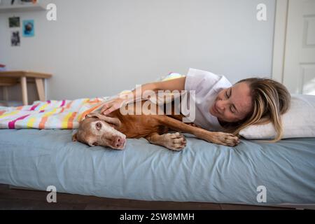 Lachende, entspannte Frau, die mit Hund Magyar Vizsla auf dem Bett liegt, faul, will morgens nirgendwo hin Stockfoto