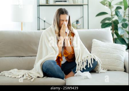Kranke junge kaukasische Frau mit langen blonden Haaren, die auf einem gemütlichen Sofa sitzt, in eine warme Decke gehüllt und in ein Taschentuch niest. Konzept von Erkältung, Grippe, Saisonkrankheiten und Allergien Stockfoto