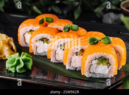 Philadelphia-Sushi-Brötchen, die mit Lachs bedeckt sind und mit Grünzeug dekoriert sind und auf schwarzem Teller serviert werden Stockfoto
