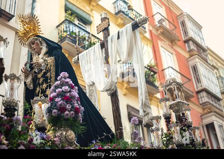 Karwoche in Malaga mit dem Thron der Jungfrau, der durch die Straßen vorrückt. Das Kreuz im Hintergrund und die Dornenkrone in ihren Händen Stockfoto