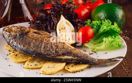 Gegrillter Fisch mit Kartoffeln und Gemüse auf weißem Teller, serviert auf Holztisch Stockfoto