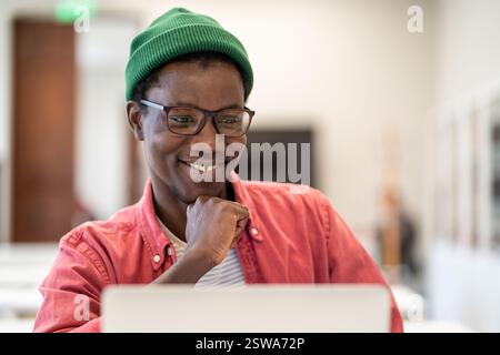 Ein fröhlicher afrikanischer Student sitzt in der Bibliothek und sieht sich den Laptop-Bildschirm an und genießt E-Learning Stockfoto
