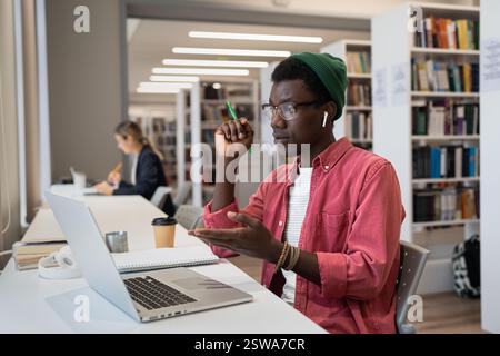 Fokussierter afroamerikanischer Mann sitzt am Tisch mit Laptop in der öffentlichen Bibliothek und spricht per Videoanruf Stockfoto