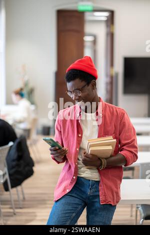 Schwarzer Student, der in sozialen Medien auf einem Smartphone chattet, während er in der Universitätsbibliothek lernt Stockfoto