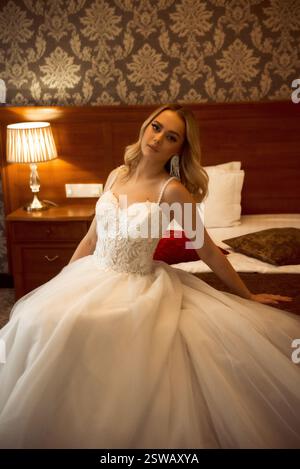 Blonde Braut sitzt in weißem Kleid auf dem Bett in stilvoller Umgebung. Stockfoto