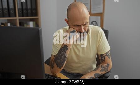 Hübscher hispanischer Mann mit Bart und Tätowierungen, der besinnlich in einem Bürozimmer mit einem Computer sitzt Stockfoto