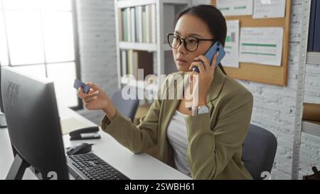 Junge Frau, die am Telefon telefoniert, während sie ihre Kreditkarte auf dem Schreibtisch des Büros hält, vermittelt eine geschäftliche, professionelle und fokussierte Atmosphäre in einem conte Stockfoto