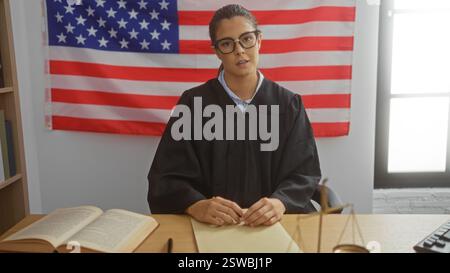 Eine Richterin sitzt in einem Gerichtssaal mit einer Brille und einer amerikanischen Flagge hinter ihr und einem Gesetzbuch auf dem Schreibtisch Stockfoto