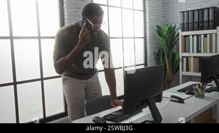 Junger Mann, der in einem modernen Büro mit Smartphone spricht, während er am Schreibtisch am großen Fenster mit Aktenschränken und Computerausstattung in hellen Professi steht Stockfoto