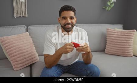 Junger Mann, der in einem Wohnzimmer sitzt und eine rote Tasse mit einem Lächeln hält, umgeben von Kissen und Wohnkultur Stockfoto
