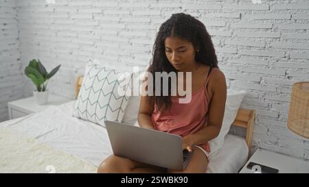 Junge Frau mit Laptop auf dem Bett in einem gemütlichen Schlafzimmer mit weißen Ziegelwänden und Pflanzen, die eine friedliche Atmosphäre schaffen Stockfoto