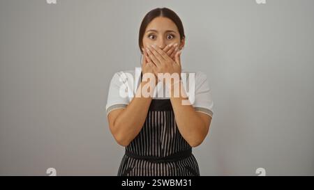 Junge Frau mit brünetten Haaren, die überraschend über isoliertem weißem Hintergrund ihren Mund bedeckt, mit weißem Hemd und gestreifter Schürze Stockfoto