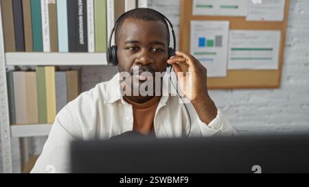Hübscher junger Mann, der in einem Büro arbeitet und ein Headset trägt, während er sich auf seinen Computerbildschirm konzentriert, mit Bücherregalen im Hintergrund Stockfoto