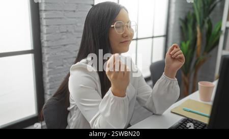Frau, die am Schreibtisch in einem modernen Büro meditiert, zeigt einen ruhigen, friedlichen Ausdruck mit Brille, geschlossenen Augen und Pflanzen, die im Hintergrund sichtbar sind Stockfoto