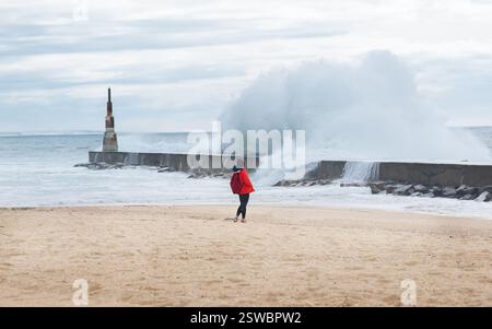 Einsame Backpackerin in roter Softshell-Jacke, die den Wintersturm genießt. Atlantische Wellen stürzen auf Breakwater mit Leuchtturm auf Praia da Aguda Sand Stockfoto