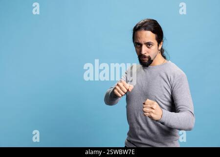 Aggressiver starker Mensch, der Fäuste sammelt und sich auf den Kampf gegen blauen Hintergrund vorbereitet, zeigt seine Stärke und Entschlossenheit für eine körperliche Herausforderung. Der Mann bereitet sich auf die Konfrontation vor. Stockfoto