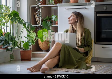Nachdenkliche Frau mit Laptop, die auf das Fenster blickt, Gefühl von Energiemangel, sitzen zu Hause mit Pflanzen auf dem Boden Stockfoto