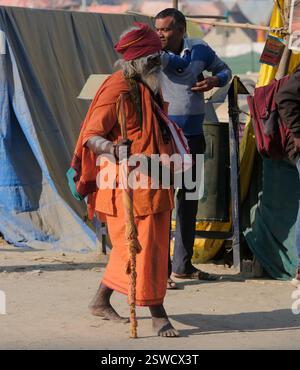Ein älterer Mann in lebendiger traditioneller Kleidung spaziert mit einem Holzpersonal durch die Straße Maha Kumbh Mela in Prayagraj und verkörpert dabei den kulturellen Geist. Stockfoto