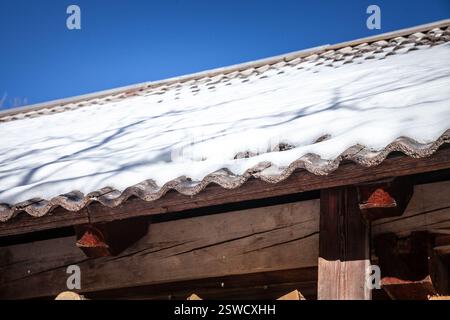 Schnee bedeckt ein rustikales Ziegeldach unter klarem Winterhimmel in Slowenien. Die frostige Schicht unterstreicht die örtlichen Bautraditionen und Isolierbedürfnisse. Stockfoto