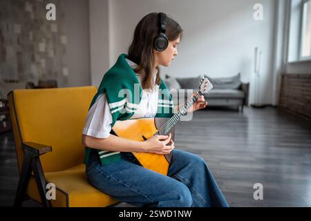 Eine fokussierte Frau spielt Balalaika, fühlt Musik zu Hause über Kopfhörer, übt das Zupfen von Saiten Stockfoto