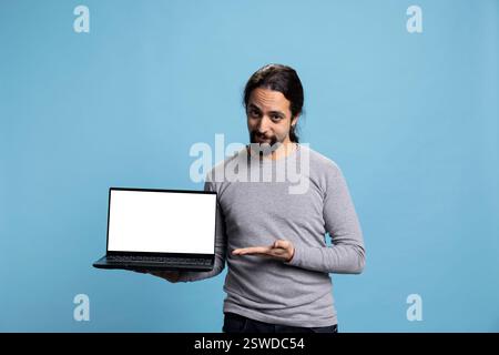 Person aus dem Nahen Osten, die auf ein Gerät mit einem weißen, isolierten Bildschirm auf der Kamera zeigt und einen Laptop hält, um einen Kopierraum für das Modell vor blauem Hintergrund zu präsentieren. Junger Erwachsener mit Freizeitkleidung im Studio. Stockfoto