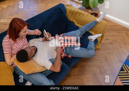 Junges, ethnisches Paar, das sich am Wochenende zu Hause mit dem Smartphone ausruht Stockfoto