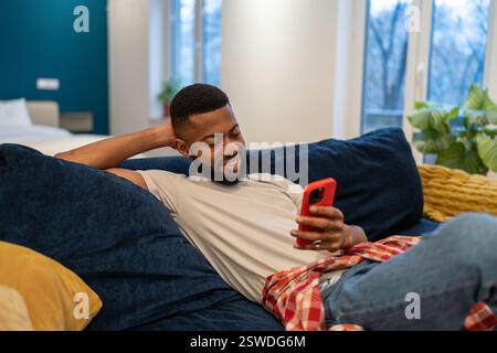 Ein entspannter lächelnder afroamerikanischer Mann sitzt auf der Couch und sieht zu Hause auf ein Smartphone. Stockfoto