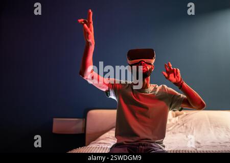 Junger afroamerikanischer Mann, der unter rotem Neonlicht im Bett sitzt und ein futuristisches metaverse VR-Headset verwendet Stockfoto