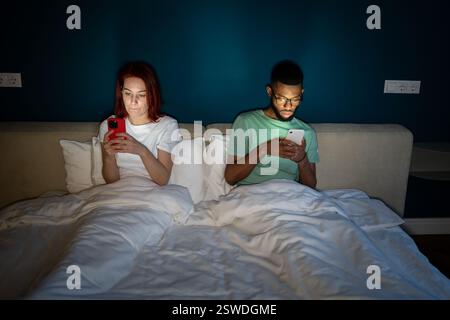 Ein junges, multiethnisches Paar, das im Bett liegt und sein Smartphone benutzt und nicht spricht Stockfoto