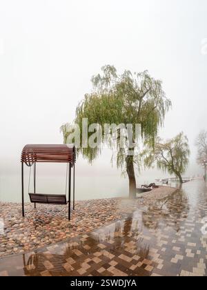 Nebelige Regenlandschaft mit modernem Park in Zheleznovodsk mit einer hölzernen Schaukelbank und einem Weidenbaum unter dem See. Kaukasus, Stockfoto