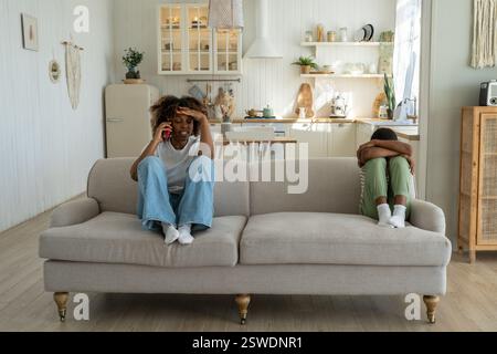 Verärgerte afroamerikanische Mutter, die mit einem beleidigten Sohn auf dem Sofa sitzt und telefoniert Stockfoto