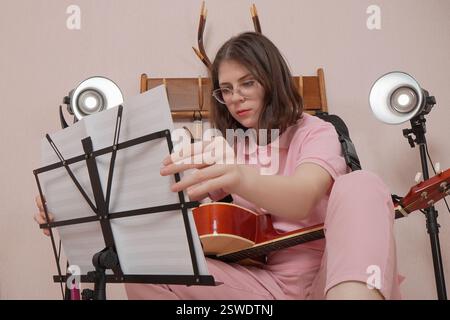 Junge Frau mit Brille und Gitarre legt Noten auf dem Notenständer und bereitet sich darauf vor, akustische klassische Gitarre im Heimstudio zu spielen. Frau des Kaukasiens Stockfoto