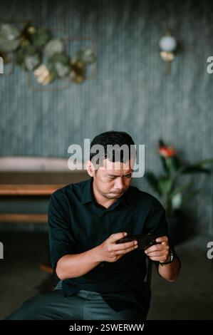 Ein Mann mit kurzen dunklen Haaren, konzentriert und nachdenklich, ist in einem Innenbereich auf seinem Smartphone-Bildschirm vertieft. Er trägt ein dunkles Hemd und eine graue Pfanne Stockfoto