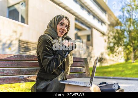 Arabische junge Geschäftsfrau, die eine Sprachnachricht mit Mobilgerät sendet und mit einem Laptop arbeitet, der draußen auf einer Bank sitzt Stockfoto