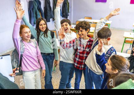 Gruppe fröhlicher Grundschüler, die Hände heben und aktiv an einem Unterricht im Klassenzimmer teilnehmen Stockfoto