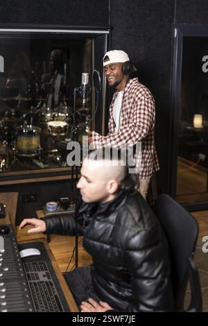 Junger Künstler, der in das Mikrofon singt, während Tontechniker Musik auf der Audiokonsole in einem professionellen Aufnahmestudio mischt Stockfoto