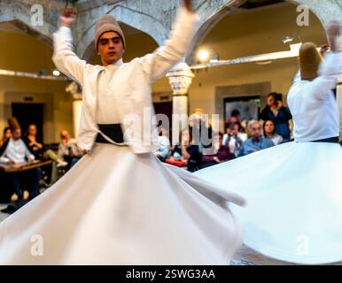 Ein wirbelnder Derwisch spielt einen traditionellen Tanz in einer kulturellen Umgebung mit fließenden weißen Kleidungsstücken und einem ruhigen Ausdruck. Das Publikum sieht zu Stockfoto