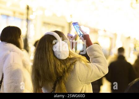 Eine Frau mit Wintergehörschützern fotografiert den lebhaften Weihnachtsmarkt in Craiova bei Nacht mit festlichen Lichtern, fröhlichem Dekor und Romanias Cheri Stockfoto