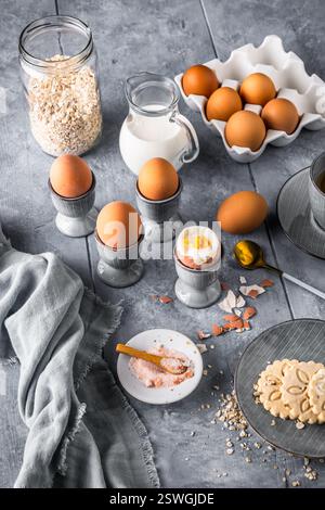 Gekochte braune Eier in grauen Eierbecher und andere Frühstückszutaten auf grauem Hintergrund, Vorbereitung zum Frühstück, vertikal Stockfoto