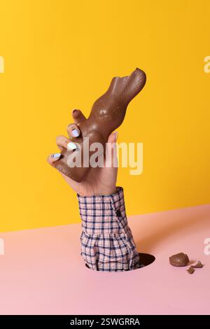 Eine tätowierte Hand mit pastellfarbenen Nägeln hält einen gebissenen Schokoladen-Osterhasen vor einem leuchtend gelben Hintergrund mit gebrochenen Schokoladenstückchen Stockfoto