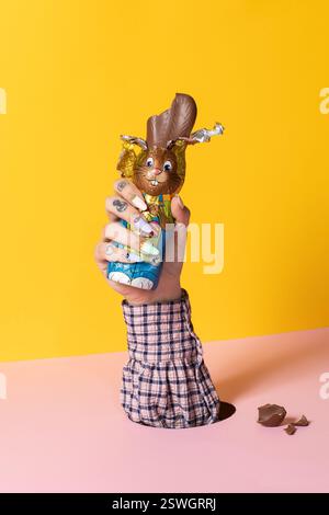 Eine tätowierte Hand mit pastellfarbenen Nägeln hält einen gebissenen Schokoladen-Osterhasen vor einem leuchtend gelben Hintergrund mit gebrochenen Schokoladenstückchen Stockfoto