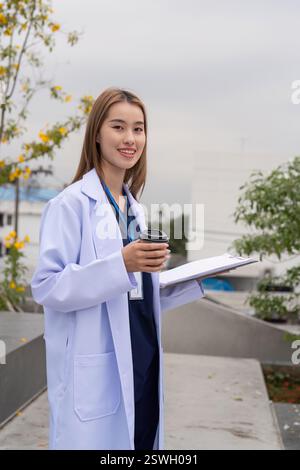Selbstbewusste junge Ärztin mit Kaffee und Tablet, lächelnd im Freien, repräsentiert moderne Gesundheitspraktiken. Stockfoto