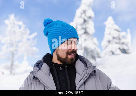 Stilvoller bärtiger Mann vor schneebedeckten Bäumen und hellblauem Himmel Stockfoto